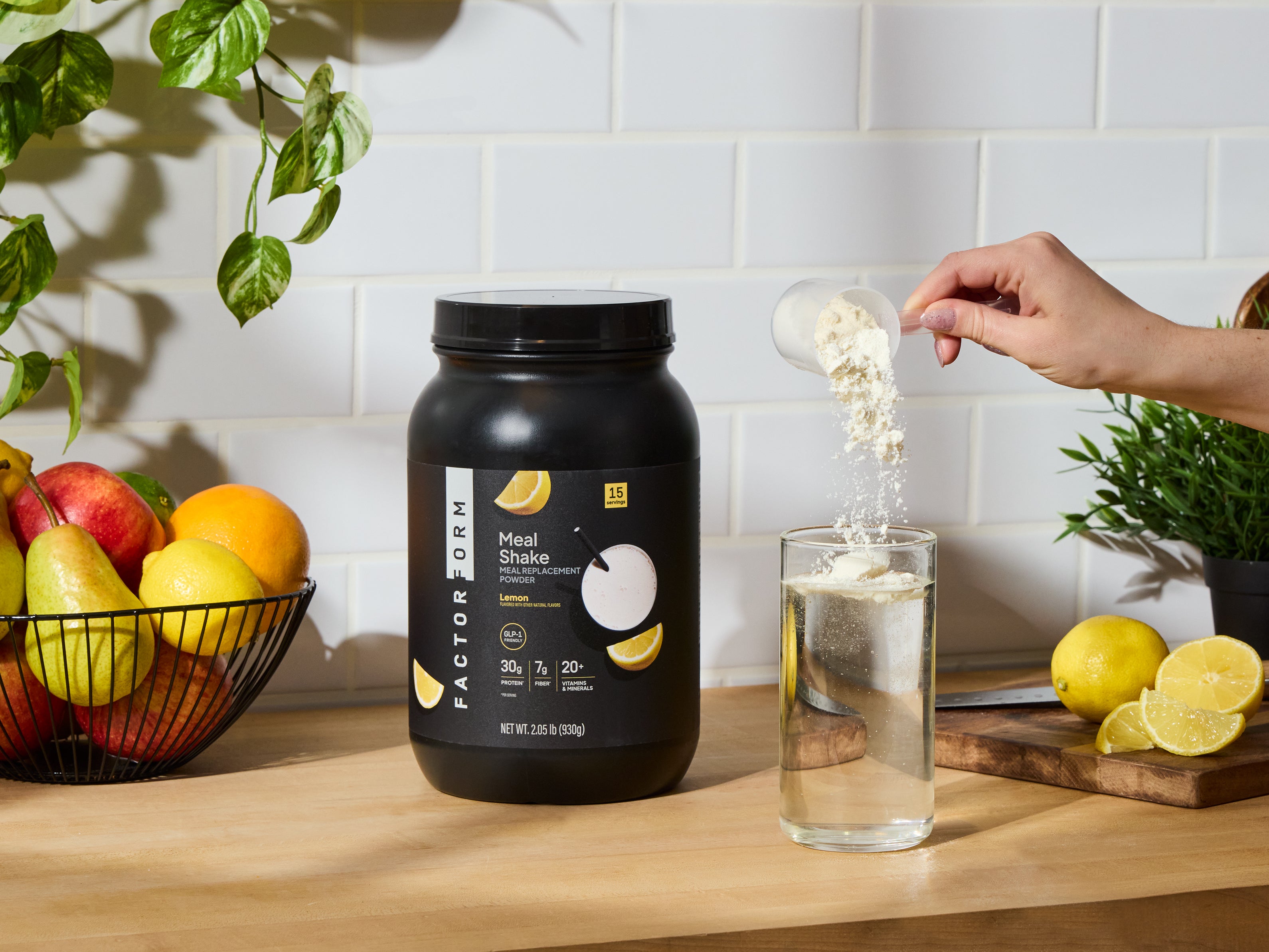 Person pouring meal shake powder from a container into a glass of water on a kitchen counter with fruits and lemons.